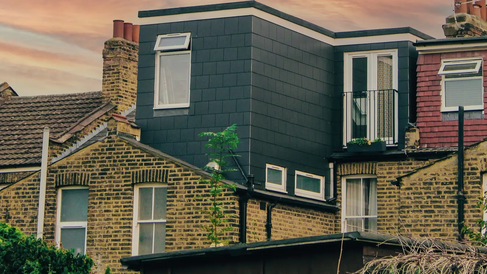 A rear dormer clad in dark tiles on the roof of a London brick terrace, with a side window and a small door with a railing, seen at sunset